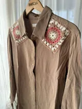 Camisa tostada de manga larga con cuello clásico decorado con apliques de crochet en tonos marrones, blancos y verdes, cierre frontal de botones y diseño holgado.