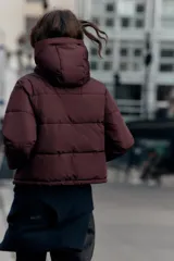 Campera puffer acolchada de color negro, con cuello envolvente, capucha integrada, manga larga, bolsillos laterales ocultos y bajo con elástico. Cierre frontal con botones a presión ocultos por solapa.