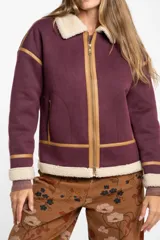 Campera tipo gamulán de color bordó, con interior y cuello de corderito sintético en tono crema. Presenta detalles de ribetes en cuero sintético color suela, cierre frontal metálico y bolsillos laterales.