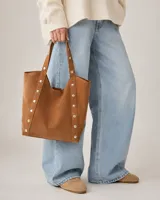 Cartera tipo tote de símil gamuza color camel con diseño amplio, correas de hombro integradas y detalles de tachas metálicas en los laterales.