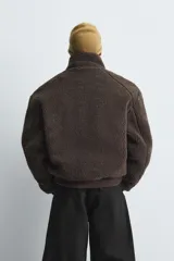 Cazadora de corte cropped confeccionada en tejido efecto borreguillo color marrón oscuro. Presenta cuello subido, cierre frontal con cremallera, bolsillos laterales de plastrón y acabados en rib en puños y cintura.