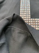 Buzo canguro negro con capucha, corte regular fit, de manga larga, con gran diseño de cruz frontal elaborado con tachas metálicas plateadas y tachas en el borde de la capucha.