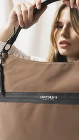 Cartera tipo hobo de tamaño grande en color beige, con detalles en negro. Presenta una franja horizontal negra con el logo de la marca, asa corta de hombro y correa larga desmontable. Incluye bolsillo frontal con cierre oculto y bolsillo trasero.