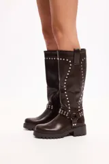 Bota de caña media en cuero sintético negro, con suela track, cierre lateral y detalle de tachas metálicas en la parte superior y en una tira decorativa sobre el tobillo.