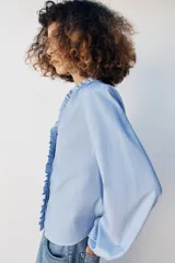 Blusa celeste de popelín, con cuello subido y escote de pico, manga larga abullonada terminada en elástico, y detalles de volantes en el cuello y la botonadura frontal con cierre de botones ocultos.
