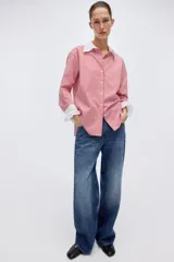 Camisa oversized de popelina de algodón con diseño a rayas verticales rojas y blancas. Presenta cuello clásico y puños anchos en color blanco sólido, cierre frontal con botones y bajo redondeado con aberturas laterales.