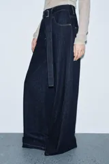 Jeans de tiro alto con pierna ancha, confeccionados en denim azul oscuro. Presentan cinco bolsillos, trabillas en la cintura y un cinturón ajustable del mismo tejido con hebilla metálica. Incluyen pespuntes marcados a tono y cierre frontal con cremallera y botón.