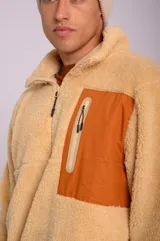 Buzo color beige de poliéster con textura tipo sherpa, cuello alto con cierre medio, bolsillo tipo parche en el pecho de color naranja con cierre vertical y puños con elástico.