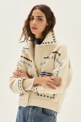 Campera de lana merino hervida en color crudo, con diseño de cuello camisero, cierre frontal de botones y bolsillos frontales. Presenta bordados artesanales con motivos geométricos en tonos marrón y azul en el frente, espalda y mangas.