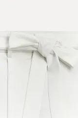 Pantalón blanco de tiro alto con trabillas, bolsillos laterales y falsos bolsillos de vivo en la espalda. Incluye cinturón lazada en el mismo tejido.