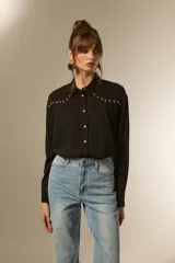 Camisa negra de viscosa con corte holgado, cuello clásico, cierre frontal de botones y detalle de tachas metálicas en la línea de los hombros.