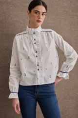 Camisa de popelina blanca con estampado de motivos deportivos, cuello alto con volados, cierre frontal con botones y mangas largas con puños elásticos y volados.