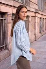 Camisa de algodón de corte holgado y diseño a rayas verticales en color celeste y blanco, con cuello camisero, cierre frontal de botones, bolsillo aplicado en el pecho y mangas largas con puños abotonados.