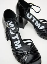 Sandalias de taco alto ancho con diseño retro, confeccionadas en charol negro. Presentan tiras delanteras múltiples entrelazadas, talón cerrado y una correa fina ajustable al tobillo con hebilla metálica.