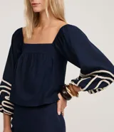 Blusa corta azul marino con escote recto estilo hombro a hombro, mangas largas abullonadas con detalle de bordado decorativo en cuerda clara.