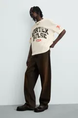 Camiseta de manga corta color crema, corte boxy fit, con cuello redondo. Presenta un estampado frontal en negro con el texto 'VORTEX PULSE' y el número '1974' en un óvalo rojo desgastado. Tiene pequeños detalles gráficos rojos en las mangas.