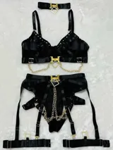 Conjunto de lencería de cuatro piezas en color negro, confeccionado con bandas elásticas y malla transparente. Incluye soutiene con aro, tiras cruzadas decoradas con tachas metálicas, cadenas y un dije romboidal dorado en el centro. La parte inferior presenta bombacha tipo tanga con tiras ajustables, arnés de piernas y portaligas incorporado con herrajes dorados.