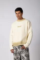 Sweater de punto acanalado en color crema, con efecto degradado en tono amarillento en el cuello y el ruedo. Presenta un diseño de cuello redondo, corte holgado y un pequeño logo bordado en el centro del pecho.
