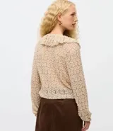 Blusa tipo bata de encaje texturizado color beige, con escote en V, mangas largas y volados aplicados en el escote. Presenta cierre frontal con botones y lazo de ajuste en la cintura.