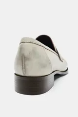 Mocasín de piel color gris claro con efecto desgastado, detalle de fruncidos en la puntera y antifaz en el empeine. Posee un taco bajo y suela oscura.