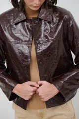 Chaqueta de efecto cuero brillante con acabado charol, cuello con solapas de piel sintética, cierre frontal con botones y bolsillos laterales.