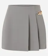 Short pollera gris de corte sastrero, confeccionado en crepe de poliéster y elastano. Presenta pliegues delanteros y un detalle de cadena metálica dorada en el lateral de la cintura.