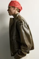 Cazadora cropped fit de color verde militar, confeccionada en tejido efecto piel. Presenta cuello subido, manga larga con puño abotonado, trabillas en los hombros, y bolsillos delanteros de plastrón con solapa. Su diseño incluye bajo elástico y cierre frontal con cremallera oculta por solapa con botones a presión.