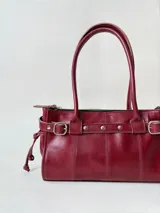 Cartera de cuero de tamaño medio con estructura rígida. Presenta una tira decorativa con tachas y hebillas metálicas en los extremos, además de lazos laterales. Cuenta con doble asa para llevar al hombro y cierre superior.