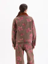 Chaqueta de gabardina color marrón con estampado floral en tonos rosados, corte recto y cierre frontal con cremallera. Presenta cuello de piel sintética en color tostado y bolsillos frontales con solapa.