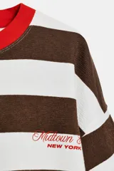 Remera de manga larga a rayas horizontales marrones y blancas, con cuello redondo rojo y detalle de texto bordado en el pecho.