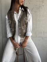 Chaleco de gamuza en color verde oliva, con diseño de bordados florales en hilo blanco y detalles de tachas metálicas. Presenta un corte clásico con cierre frontal de botones.