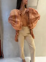 Blusa de tela liviana con efecto texturizado en tono terracota, caracterizada por sus mangas largas extremadamente amplias y abullonadas. Presenta un escote en V y un diseño fluido con terminaciones irregulares.