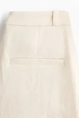 Pantalón sastrero de tiro alto en color beige, con perneras amplias y raya marcada en la parte delantera y trasera. Cuenta con cierre de cremallera, botón oculto y corchete, además de bolsillos decorativos insertados.