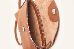 Cartera tipo sobre de cuero color marrón, con solapa frontal redondeada que presenta un detalle circular grabado. Incluye correa larga ajustable y extraíble, cierre principal y broche de seguridad.