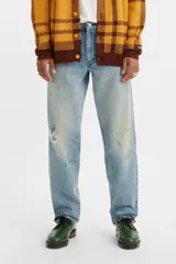 Jean de corte relajado y pierna cónica, con diseño de cinco bolsillos y cierre frontal con botones. Presenta un lavado azul claro con efecto desgastado, roturas localizadas y detalles de salpicaduras de pintura.