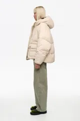 Chaqueta acolchada tipo puffer de corte globo en color beige, con cuello subido, capucha, cierre frontal con cremallera y botones de presión, y bolsillos delanteros con tapeta.