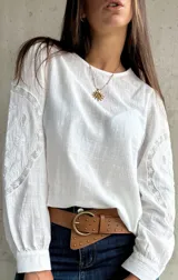 Blusa blanca de lino con mangas largas abullonadas, destacada por paneles laterales bordados con detalles de encaje. Presenta un corte holgado y cuello redondo.