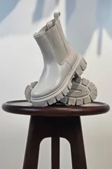 Bota tipo Chelsea de caña corta en color beige, confeccionada en material sintético con acabado liso. Presenta paneles elásticos laterales a tono y una suela track robusta y dentada que aporta altura y estilo moderno.