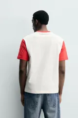 Remera de corte regular fit en color blanco con cuello redondo y mangas cortas en color rojo contrastante, y aplicación de parche bordado en el pecho.