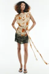 Vestido corto sin mangas de cuello redondo, con estampado tie-dye en tonos beige, naranja y verde oscuro. Presenta un lazo para atar en la cintura y cierre trasero con cremallera oculta.