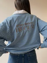 Buzo tipo chomba de corte oversize con diseño a rayas horizontales celestes y blancas. Presenta cuello camisero blanco y un bordado en la espalda con la inscripción República Oriental del Uruguay.