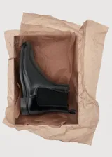 Botín tipo Chelsea de cuero vacuno color negro, con paneles laterales elásticos y tirador trasero. Presenta una silueta clásica con punta redondeada y suela plana de resina de poliuretano.