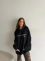 Canguro negro oversize con capucha, cordones de ajuste y bolsillo frontal tipo canguro. Presenta estampado con texto en blanco en el pecho.