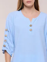 Blusa Erika de algodón con estampado de rayas en tonos azules, rosados y amarillos. Presenta cuello redondo con escote en V y botones frontales, mangas cortas decoradas con una fila de botones en los laterales y dobladillo asimétrico.