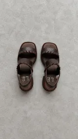 Sandalias de cuero marrón con punta cuadrada, tiras texturizadas con detalles de ojales metálicos y cordones entrelazados, y tira ajustable con velcro en el talón.