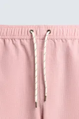 Bañador largo de hombre color rosa pálido con textura seersucker. Presenta cintura elástica ajustable con cordón blanco, bolsillos laterales y un bolsillo trasero de plastrón.