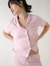Camisa de manga corta para dormir de color rosado, confeccionada en viscosa. Presenta botones frontales, cuello con solapa, bolsillo en el pecho y ribetes a tono.