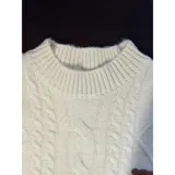 Sweater de punto color crema con diseño de trenzas y rombos, cuello redondo y mangas largas. Presenta un pequeño aplique decorativo en el pecho.