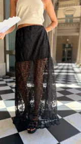 Falda larga de encaje negro con diseño de capas, presentando una sección superior opaca y una sección inferior transparente con terminación de volado en el ruedo.