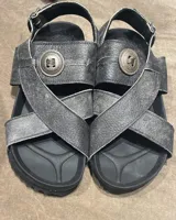 Sandalias de cuero vacuno en color gris oscuro con acabado desgastado. Presentan un diseño de tiras cruzadas con un aplique metálico circular decorativo en el centro y cierre con hebilla en el talón. Cuentan con plantilla anatómica y suela de goma negra.